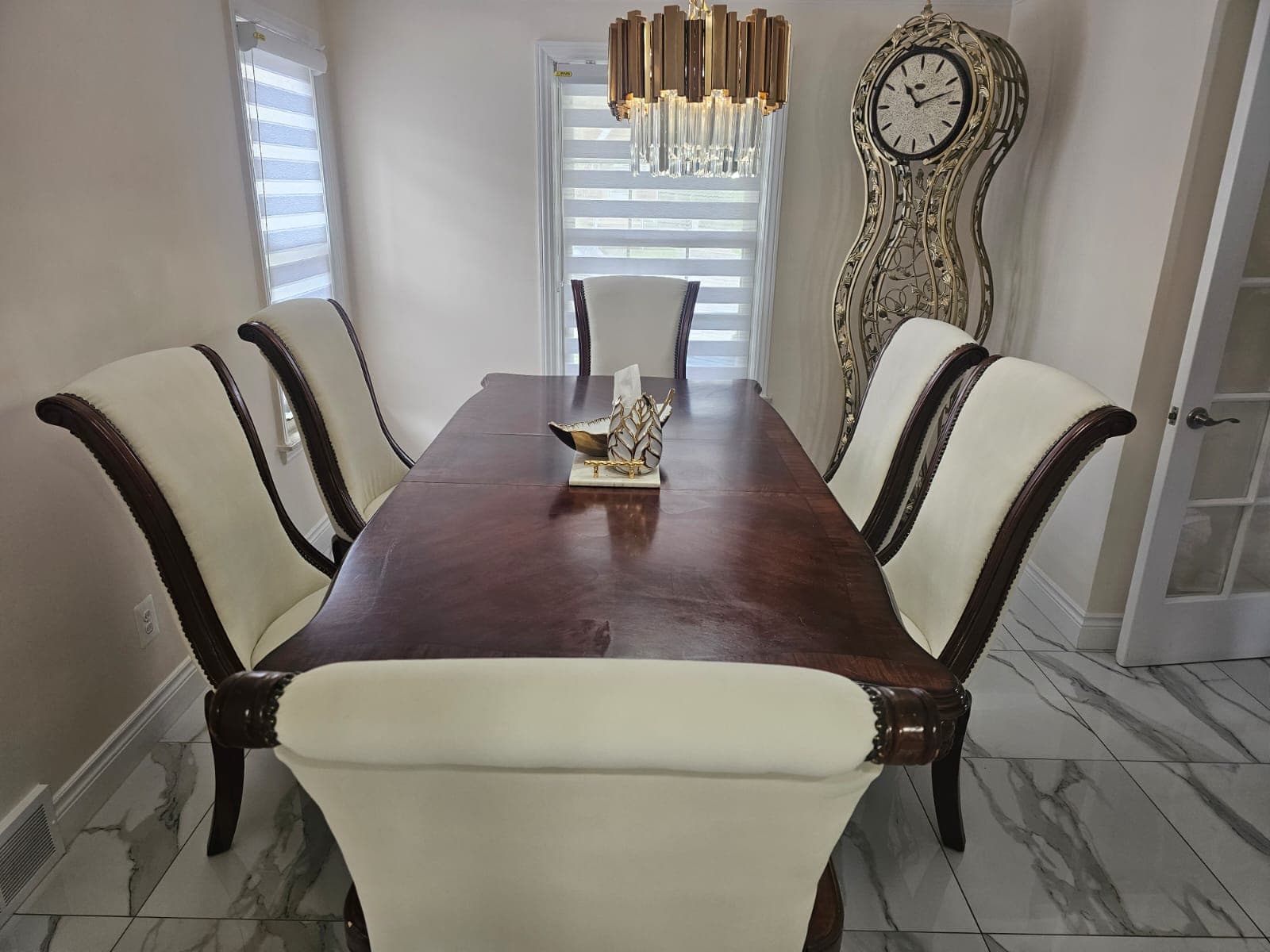 Formal dining room with dark wood table, white chairs, gold chandelier, and ornate grandfather clock.